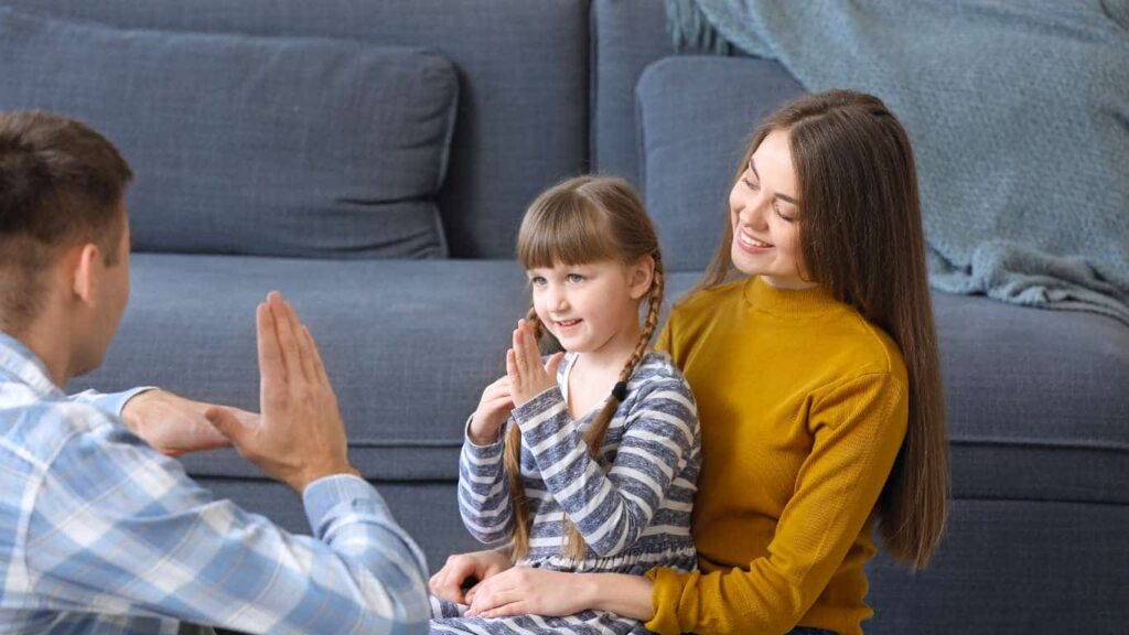 Little girl with light brown hair in bangs and two braids is sitting on her mom's lap while in front of a grey couch. She's signing to a gentleman who is sitting on the floor across from her.