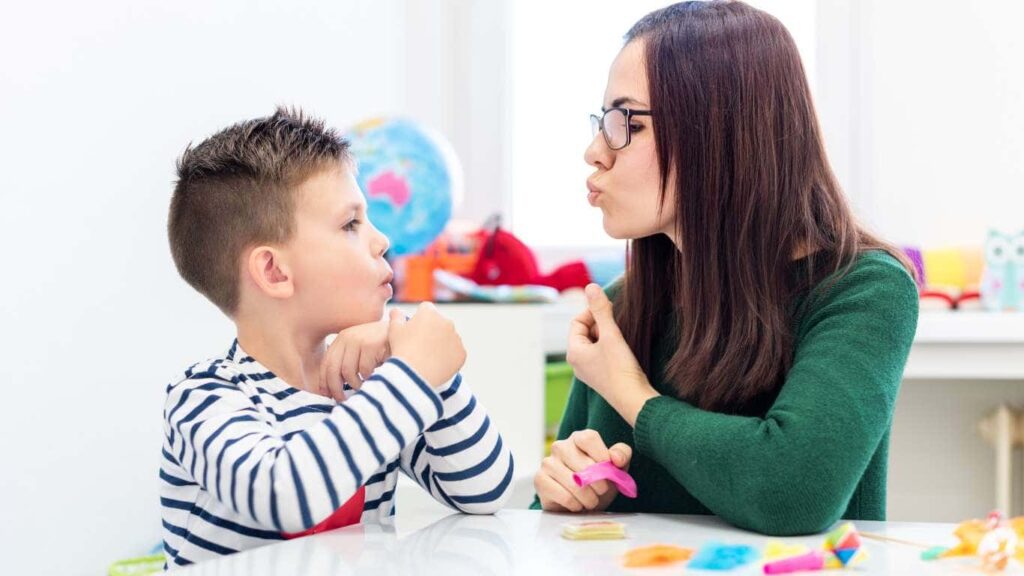 speech pathologist with long brown hair and green sweater puckering her lips to demonstrate how to make a speech sound as she faces her young child client