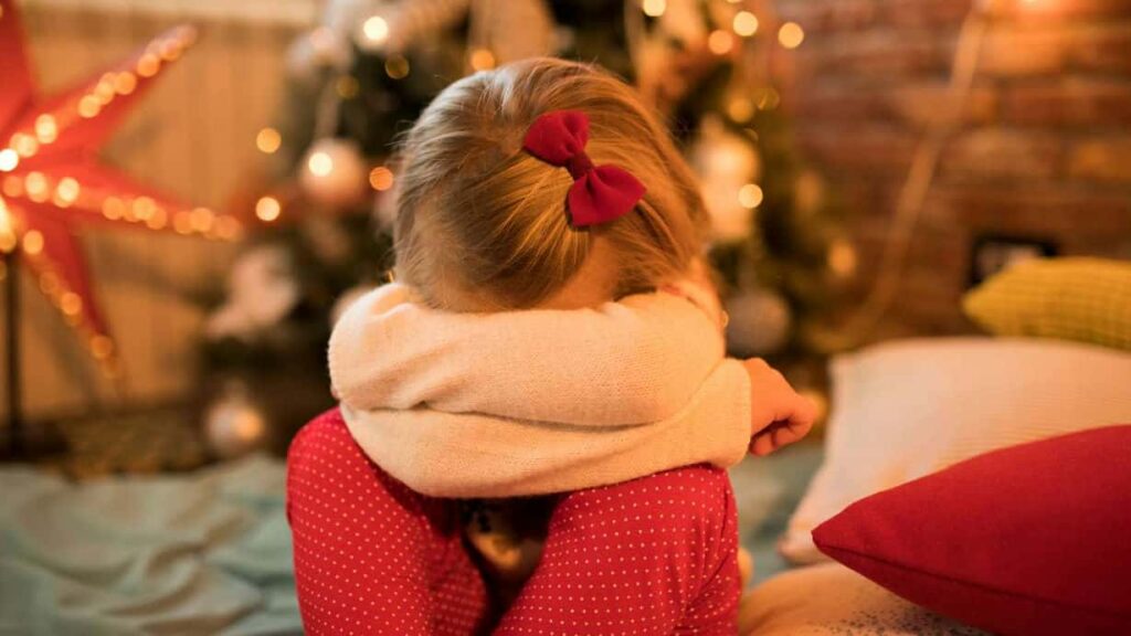 Little girl with blond hair wearing a long-sleeve white shirt and red pants. She's in the living room in front of a Christmas tree and her head is in her lap. She appears to be pouting or crying.