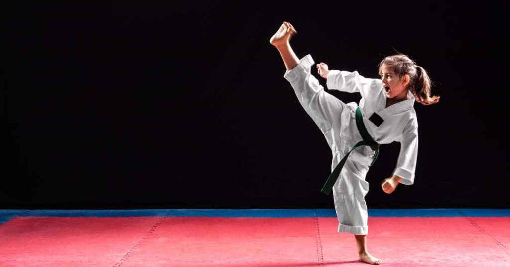 Little girl with blond ponytail wearing a white taekwondo dobak while kicking her leg in the air. She is standing on a red and blue mat with a black wall in the background.