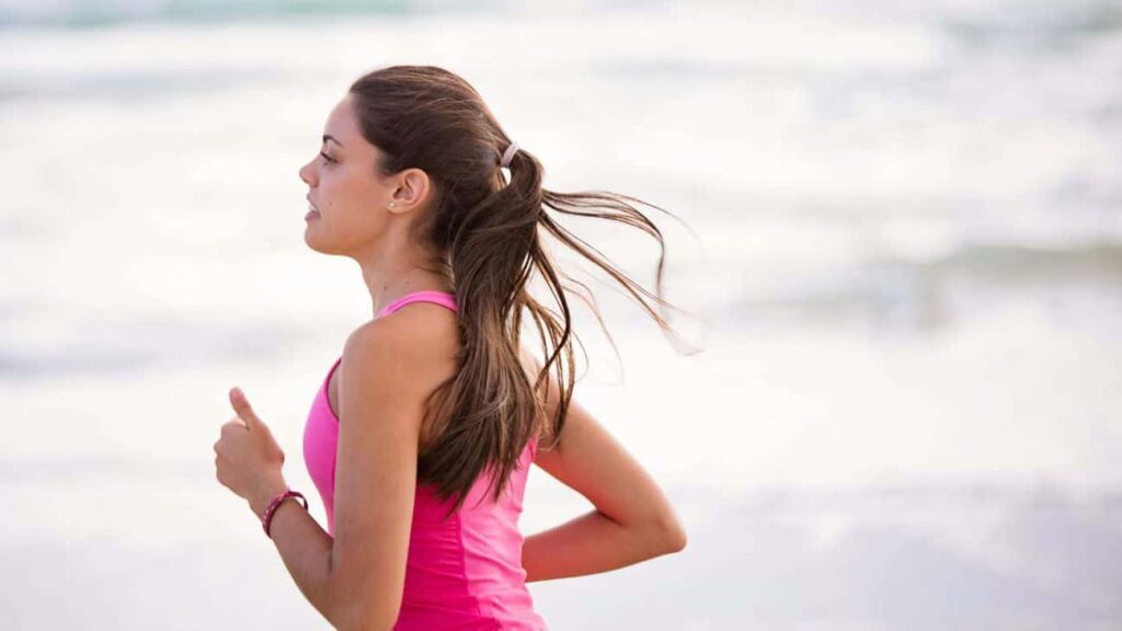 Woman with long brown hair in a half ponytail running along the beach. She's wearing a pink tank top.