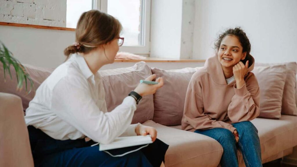 A mental health therapist sitting across from her client. They're both sitting on a tan couch looking at one another while talking.