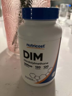 Bottle of Nutricost DIM hormone supplement. White bottle with blue cap against a pink vase on a white countertop.