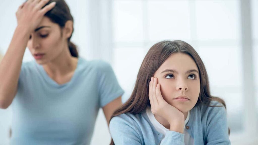Mom and teen daughter. Daughter staring off into space with an attitude. Mom's hand is on her forehead in frustration. Both mom and teenage daughter are wearing light blue shirts.