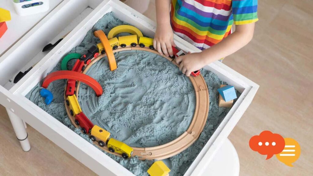 Child wearing a rainbow striped shirt playing with trains in a sandbox.