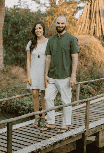 Woman with long brown hair wearing a white dress is holding her husband's hand. He is balding and has a brown beard. Both are smiling and standing on a small wooden bridge.